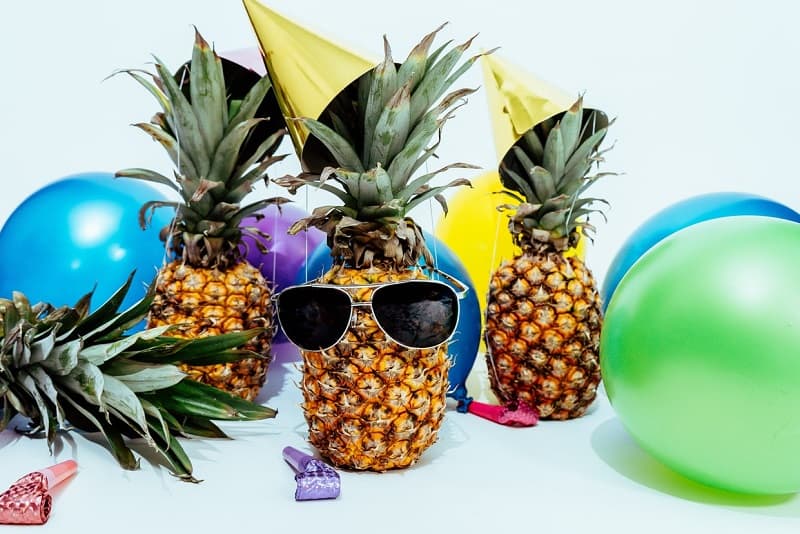 Three pineapples wearing sunglasses and party hats surrounded by balloons